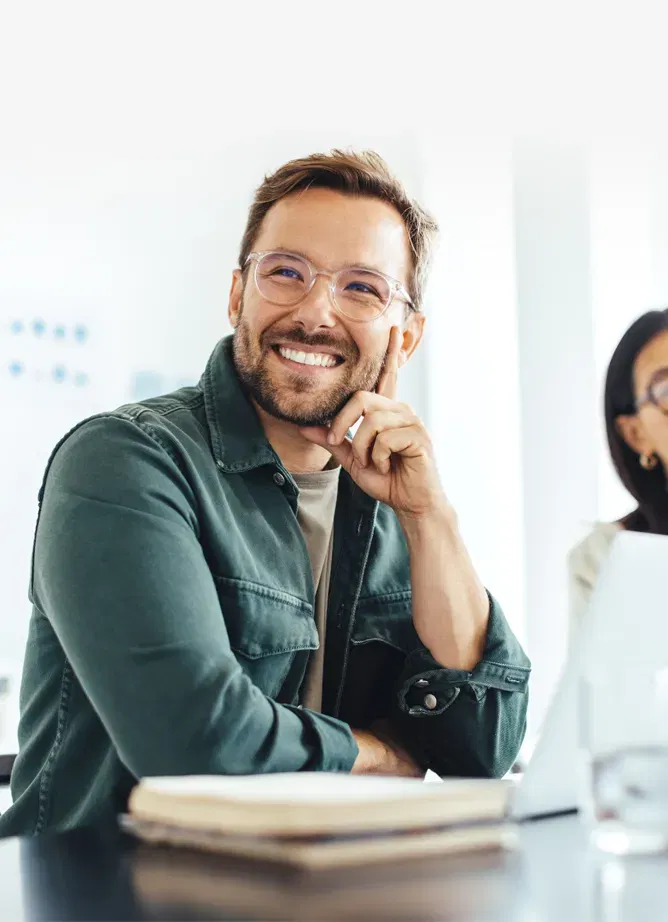 A man wearing glasses sits at a desk and smiles, next to an out of focus woman