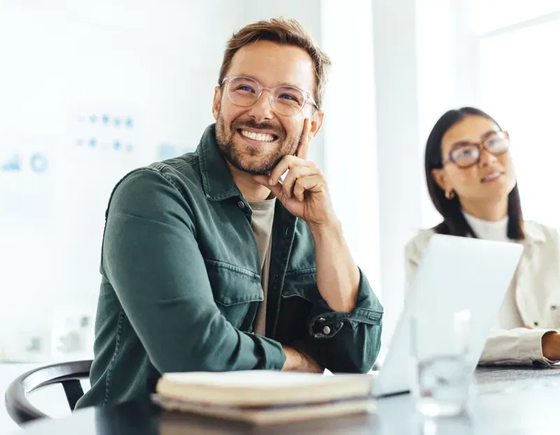 A man wearing glasses sits at a desk and smiles, next to an out of focus woman