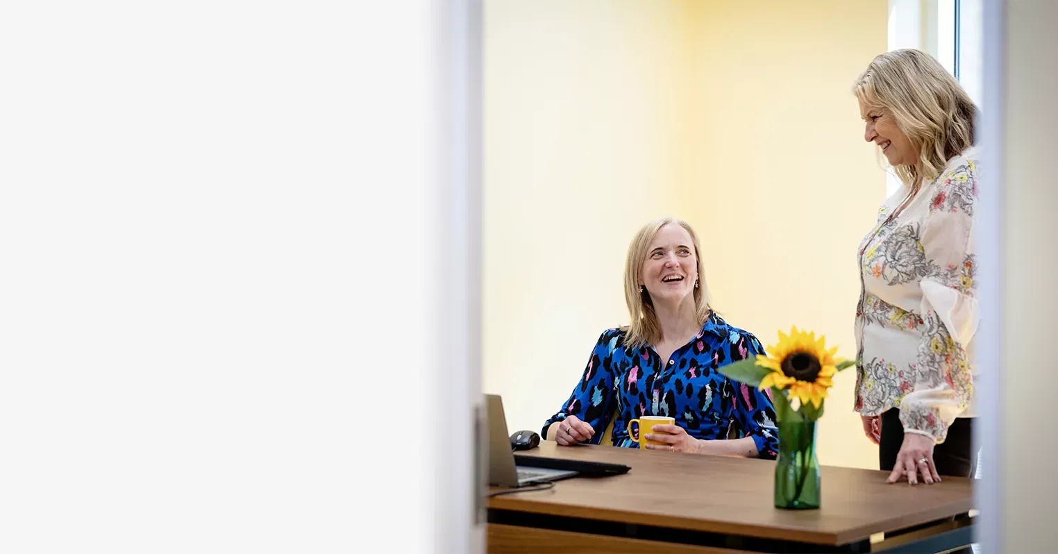 Two smiling women, one standing up, the other seat and holding a yellow mug