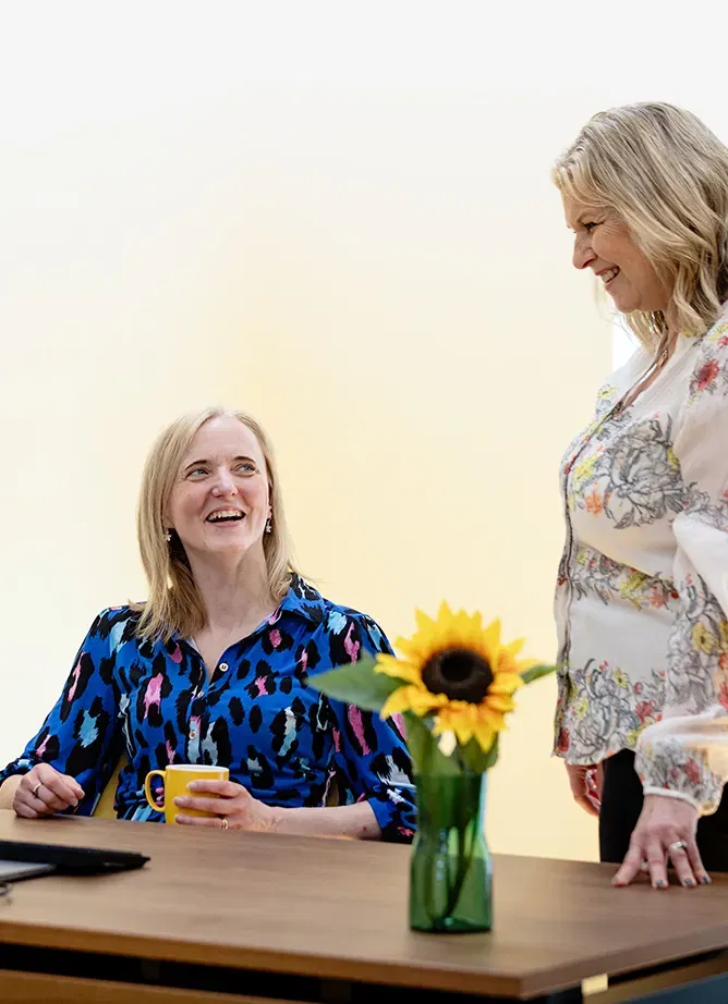 Two smiling women, one standing up, the other seat and holding a yellow mug