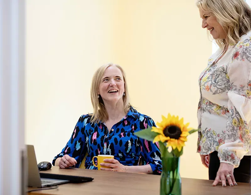 Two smiling women, one standing up, the other seat and holding a yellow mug