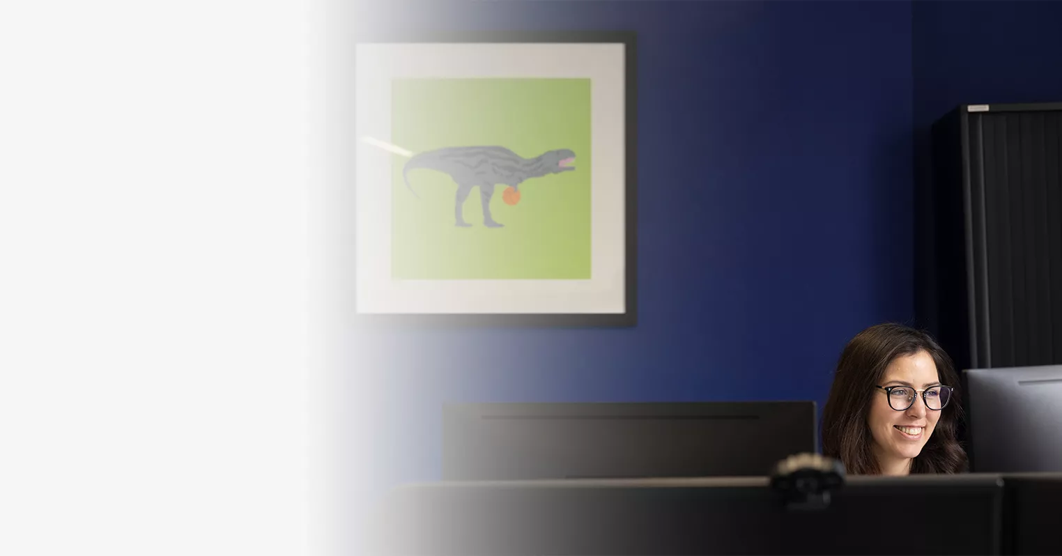 Photo of woman with glasses at desk in front of monitor. Framed wall picture of cartoon dinosaur behind her.