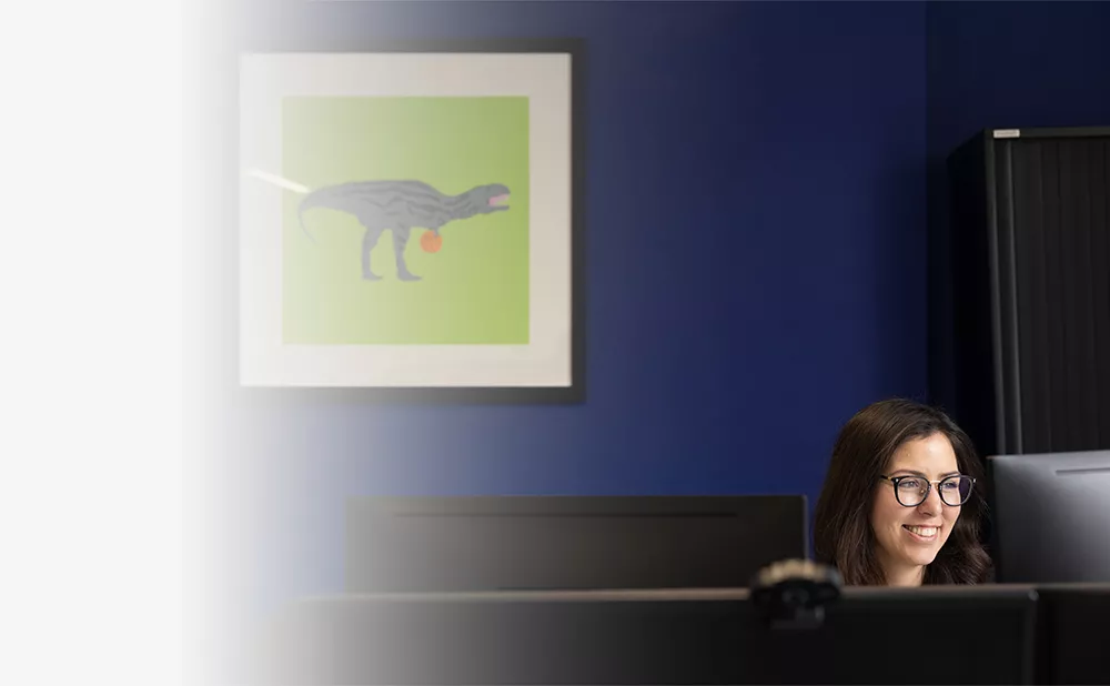 woman with glasses at desk in front of monitor. Framed wall picture of cartoon dinosaur behind her.