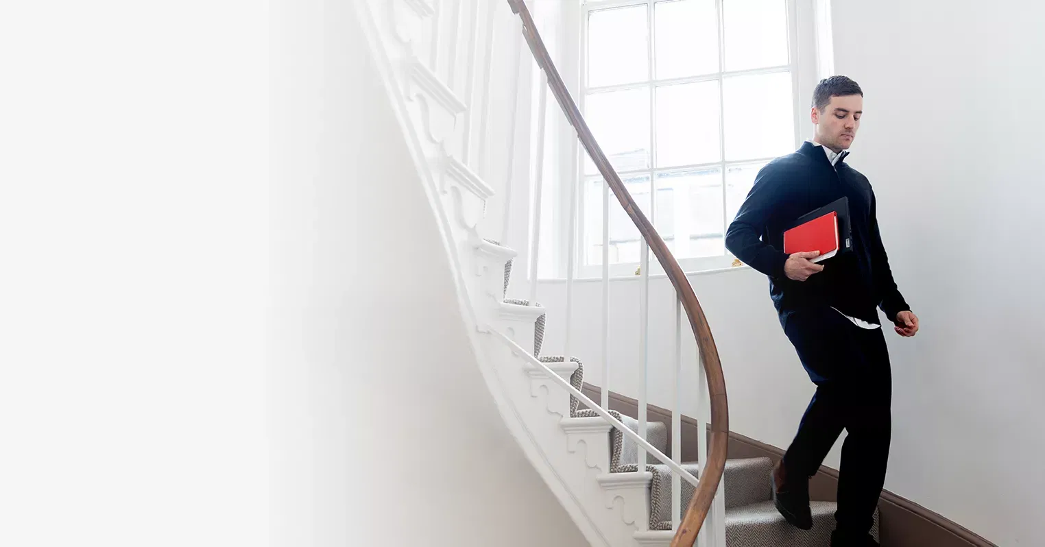 A man descends a curved staircase holding a tablet and red notebook