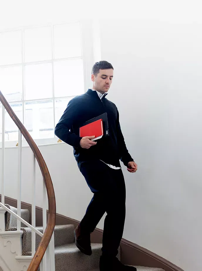 A man descends a curved staircase holding a tablet and red notebook