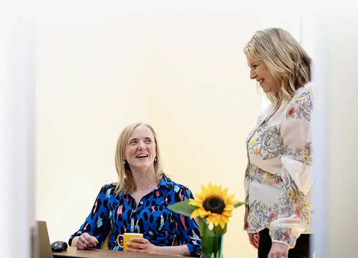 Two women, one seated and one stood, smile and laugh in an office setting